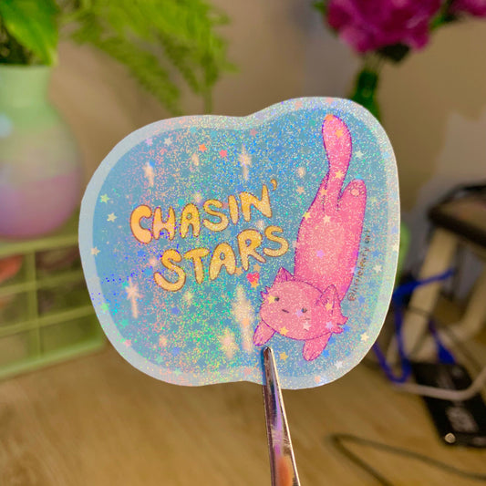 Holographic sticker with 'Chasin' Stars' text and a pink cat design held by scissors.