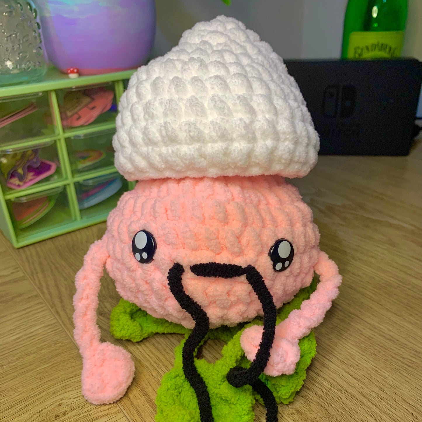 Crocheted plush toy with a white hat, pink face, and green base on a wooden surface.