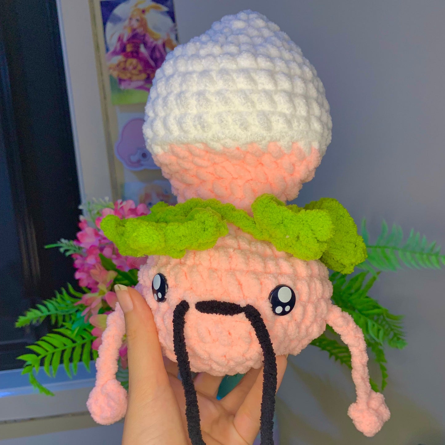 Hand holding a small plush toy with a white hat and green leaves in a casual setting.