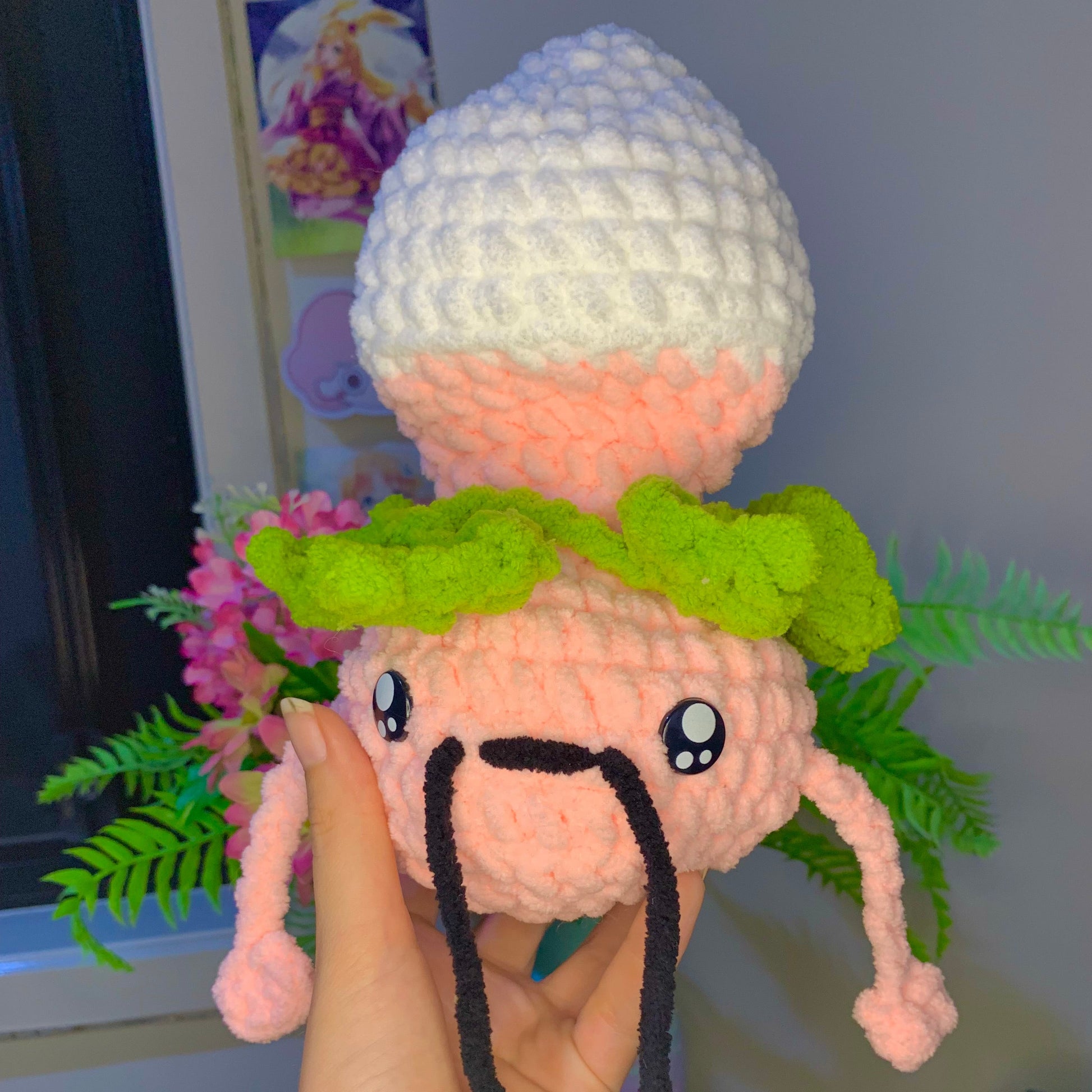 Hand holding a small plush toy with a white hat and green leaves in a casual setting.