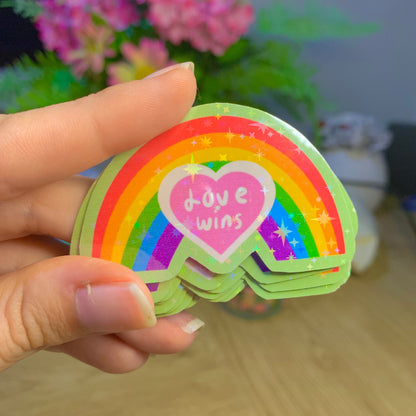 Hand holding a stack of rainbow-colored stickers with a heart and text, blurred background with flowers.