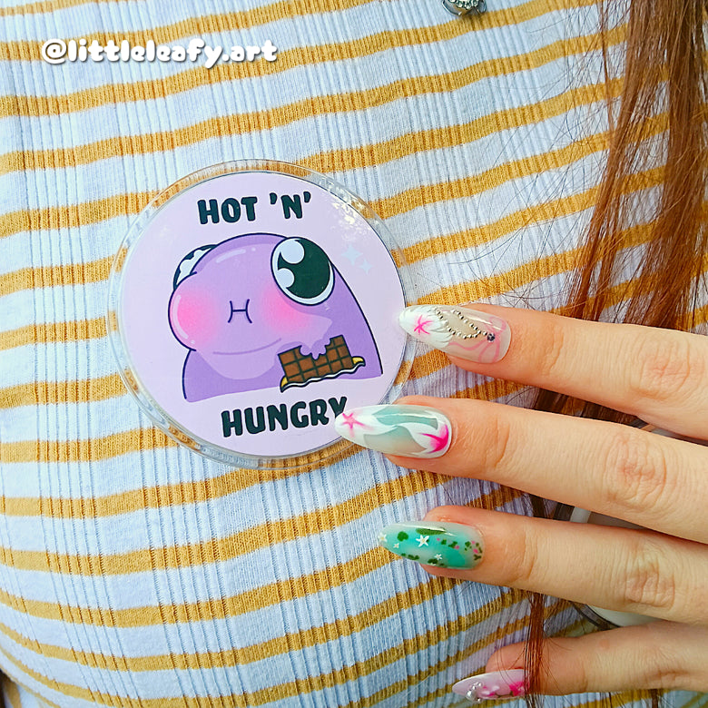 Circular badge with cartoon character and text 'Hot N' Hungry' held by a person with colorful nails on a striped background.