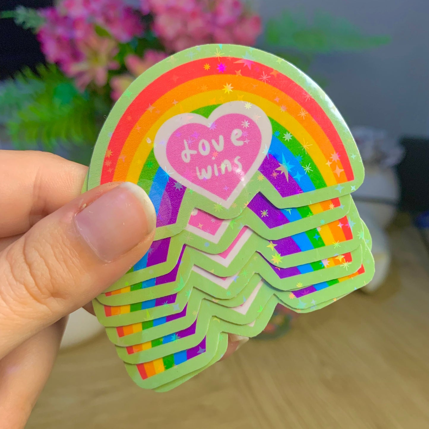Stack of rainbow-themed stickers with a heart and 'Love wins' text held by a hand, with a blurred background.