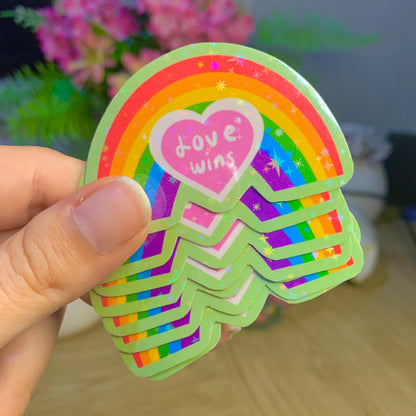 Stack of rainbow-themed stickers with a heart and 'Love wins' text held by a hand, with a blurred background.
