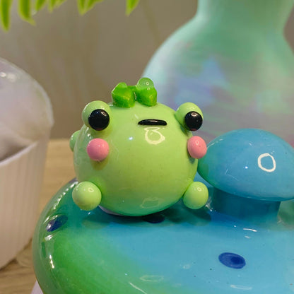 Green frog with black eyes and pink cheeks on a blue and green base