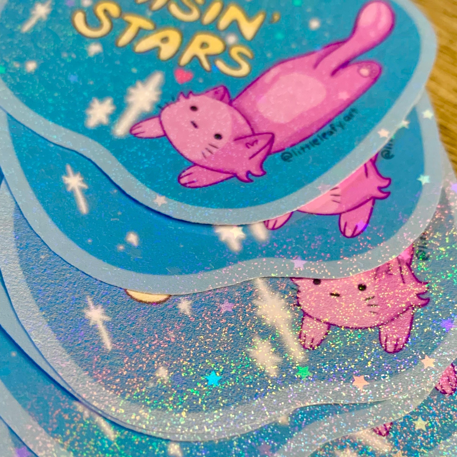 Stack of glittery stickers with a pink cat design and 'asin stars' text on a blue background.
