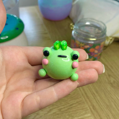 Small green frog figurine held in a hand on a wooden surface with colorful objects in the background.