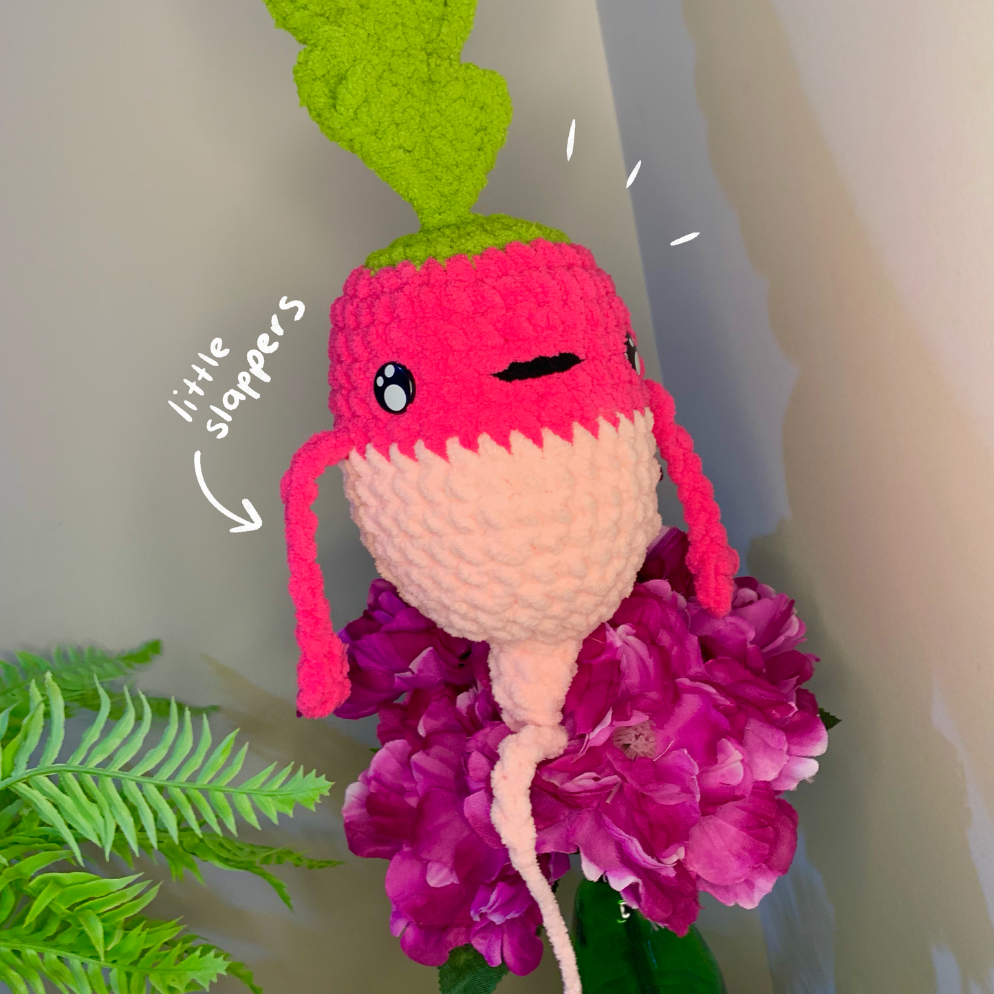 Crocheted radish character with pink top and green leaves, surrounded by purple flowers on a neutral background.