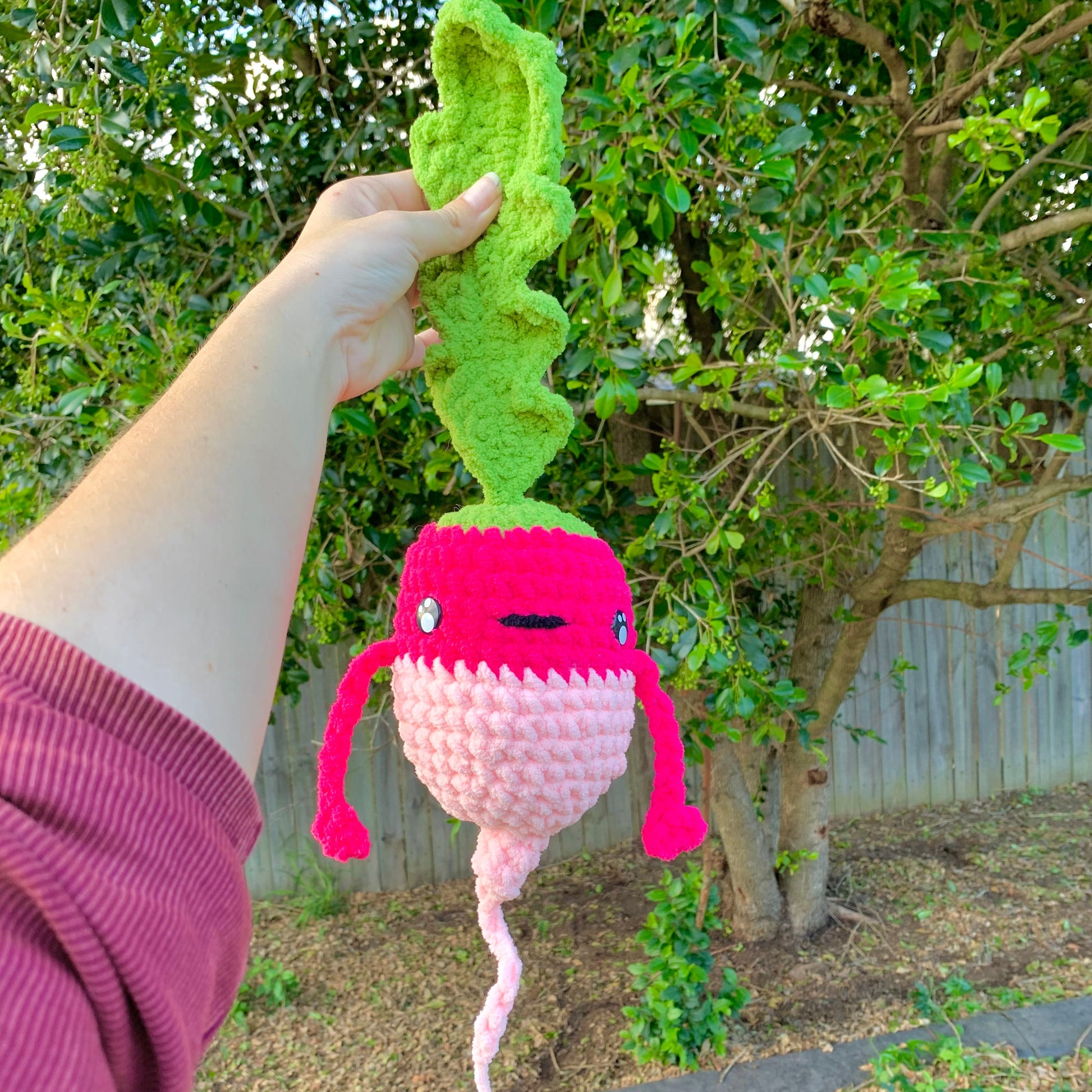 Hand holding a pink and green knitted radish-shaped toy outdoors.