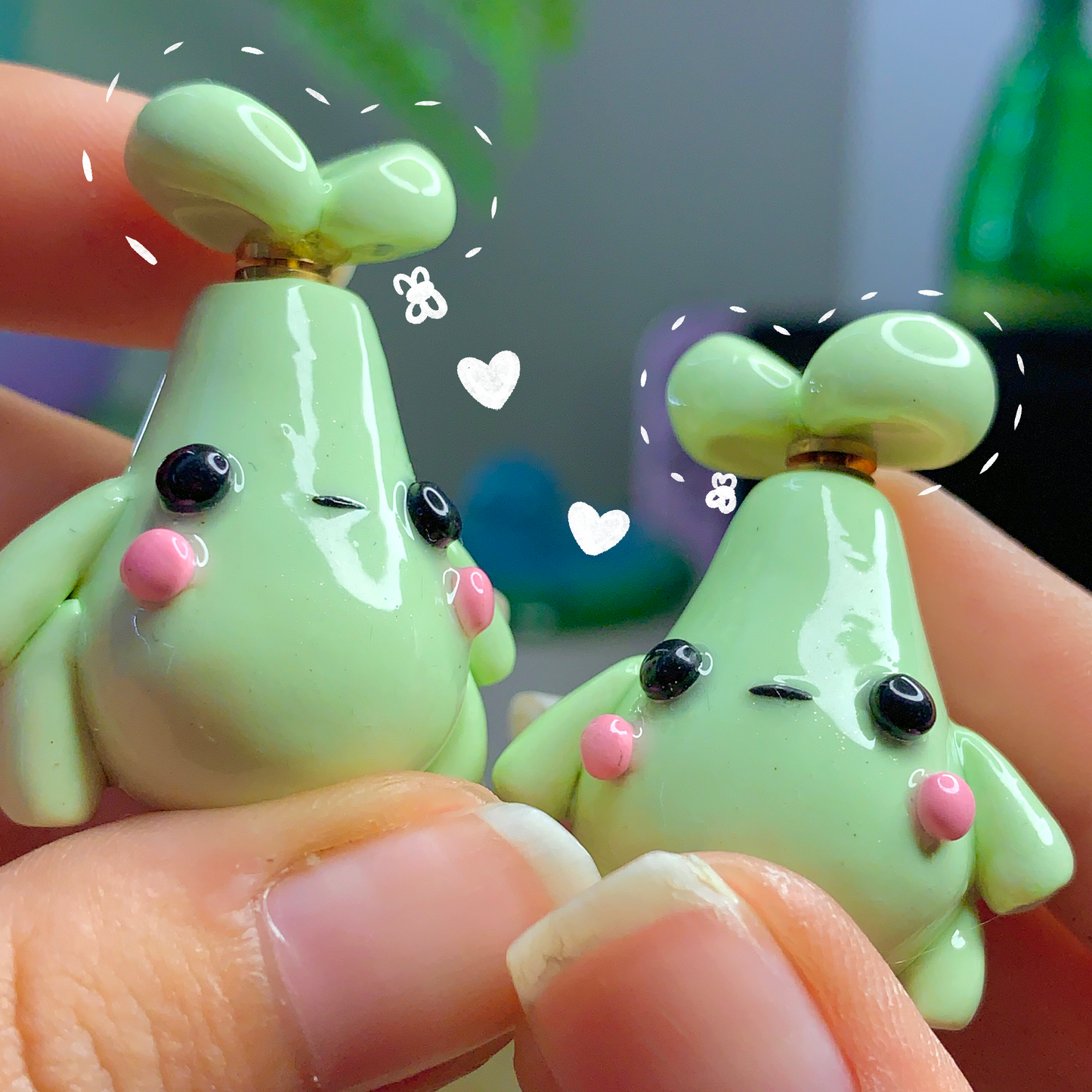 Two small green figurines with black eyes and pink cheeks held between fingers.