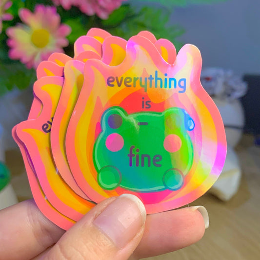 Colorful card with 'everything is fine' text held by a hand, with flowers in the background.
