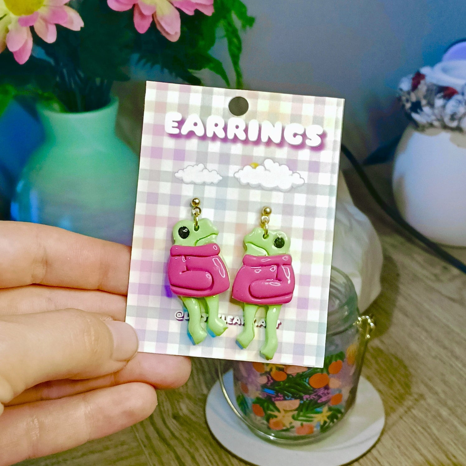 Earrings with frog design on a card held by a hand, with a colorful background.